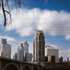 The Minneapolis skyline with the Stone Arch Bridge in the foreground and blue sky and white clouds in the background.