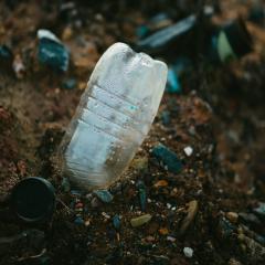 A plastic water bottle pokes out of brown soil dotted with rocks, with other plastic trash out of focus in the background.