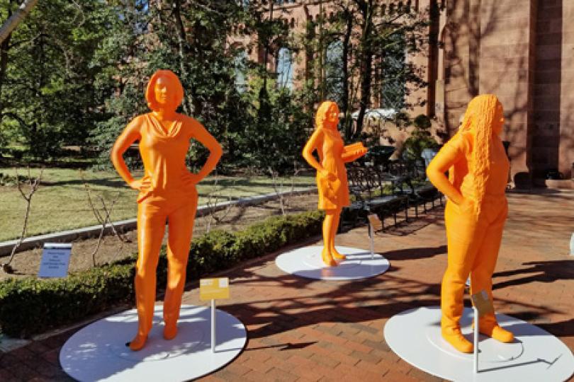 Life-sized, bright orange statues of three women, including Dr. Kimberley Miner, in a brick courtyard outside a brick Smithsonian building with trees in the background.