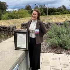 Lisa Micheli stands on an outdoor patio with a stone wall, grass field, trees and cloudy sky in the background, smiling and posing with a framed copy of her award. 