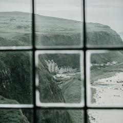 A black and white view seen through a window pane of a cliff overlooking a beach with buildings and a pier. 