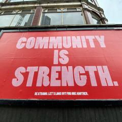 A red billboard says "COMMUNITY IS STRENGTH" in large pink letters. Smaller text beneath it reads "BE STRONG. LET'S LOOK OUT FOR ONE ANOTHER."