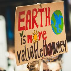 A cardboard sign is painted with the text “EARTH is more valuable than money” and a drawing of Earth. 