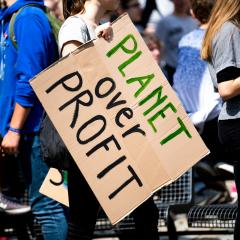 A white woman holds a cardboard sign that reads Planet over Profit in green and black text, standing in a crowd of people.