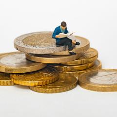  A miniature person reads a newspaper while sitting on a pile of golden coins against a white background.