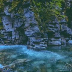 A river flows alongside gray stone cliffs with green moss growing on them. 