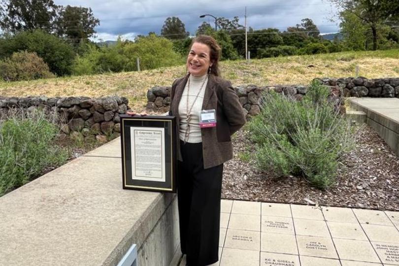 Lisa Micheli stands on an outdoor patio with a stone wall, grass field, trees and cloudy sky in the background, smiling and posing with a framed copy of her award. 