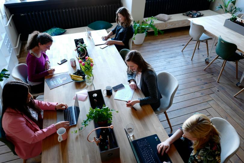 Seen from above: five women sit around a wooden table with laptops and notebooks, smiling and working together.