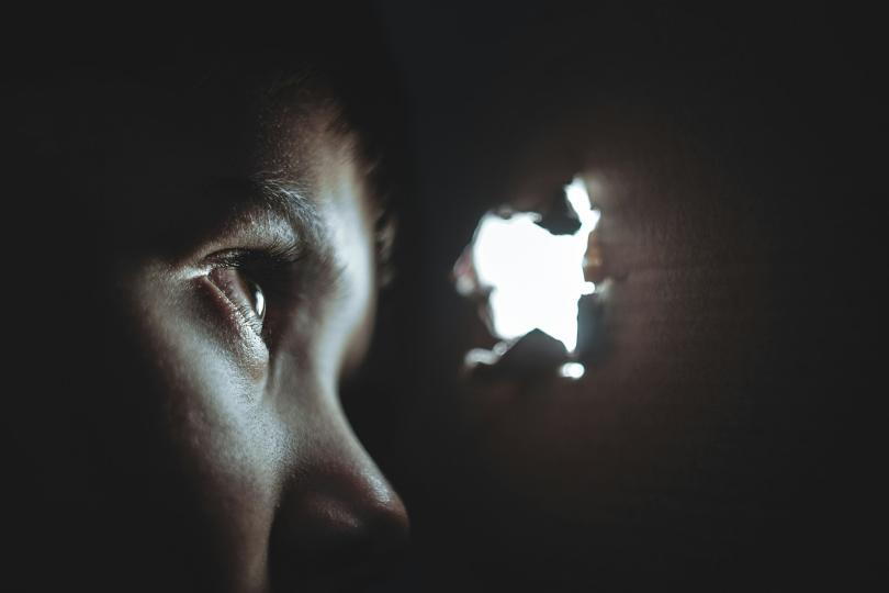 A child’s face peers at light coming through a hole into a dark space.