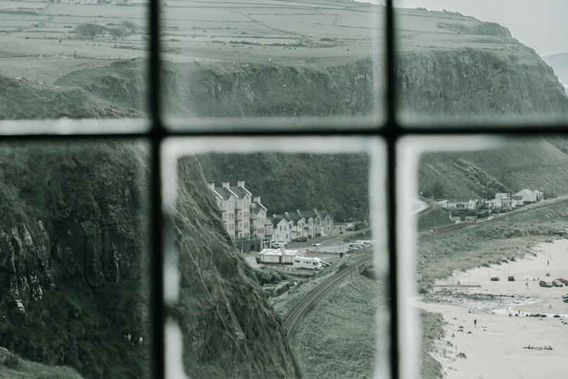 A black and white view seen through a window pane of a cliff overlooking a beach with buildings and a pier. 