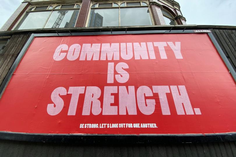 A red billboard says "COMMUNITY IS STRENGTH" in large pink letters. Smaller text beneath it reads "BE STRONG. LET'S LOOK OUT FOR ONE ANOTHER."