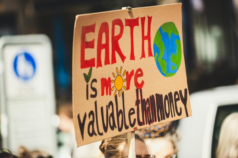 A cardboard sign is painted with the text “EARTH is more valuable than money” and a drawing of Earth. 