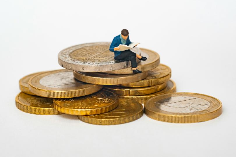  A miniature person reads a newspaper while sitting on a pile of golden coins against a white background.