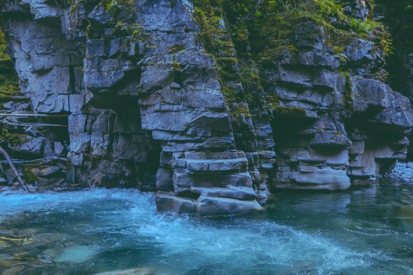 A river flows alongside gray stone cliffs with green moss growing on them. 