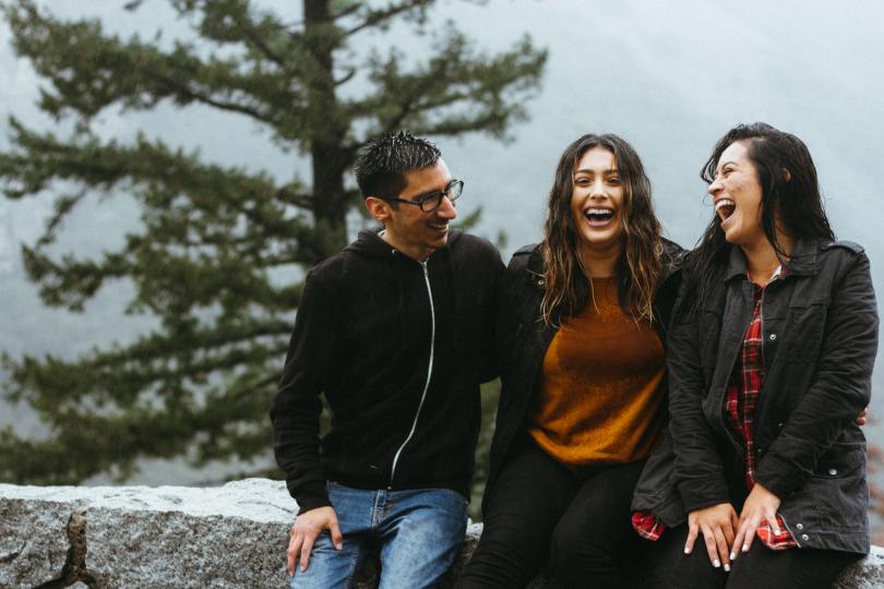 Three people sit on a concrete wall in front of trees, smiling and laughing in the rain.